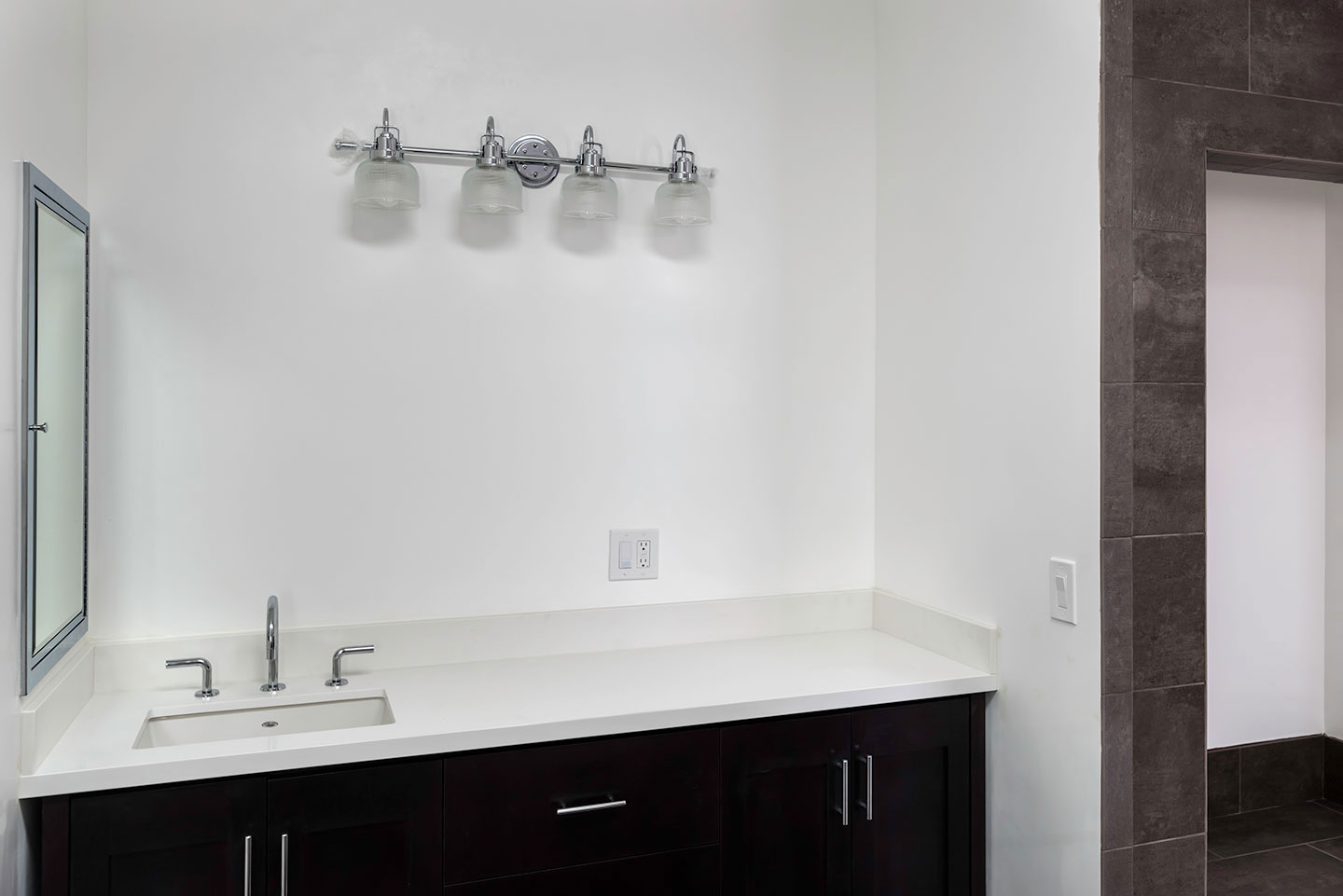 The image depicts a modern bathroom with a white sink countertop, dark cabinets, a black tile backsplash, a gray floor, and a single-handle faucet.