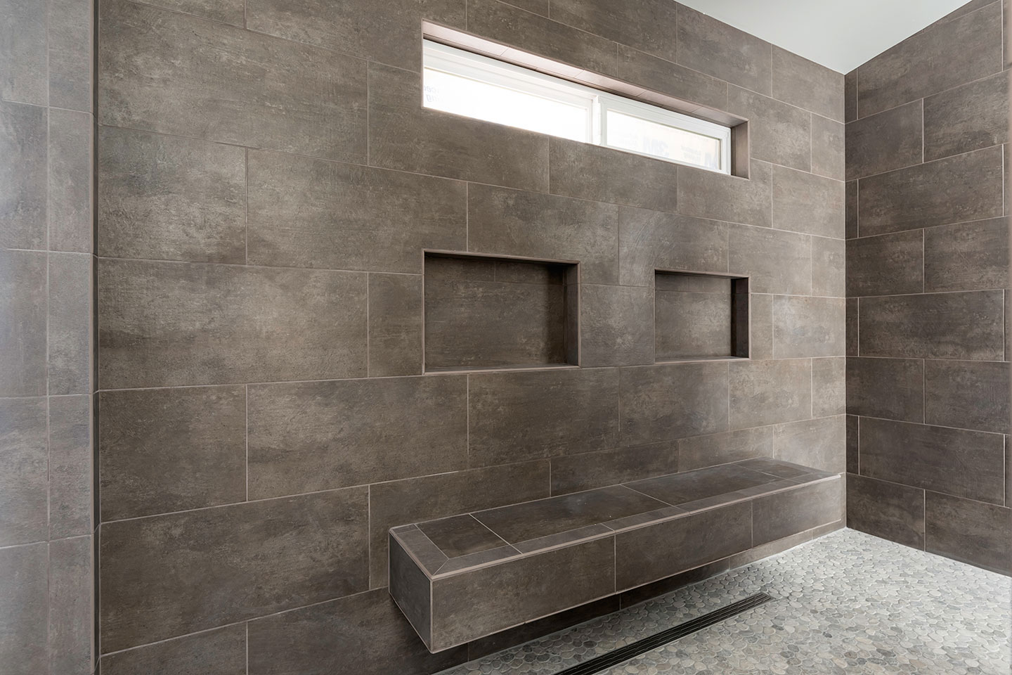 The image depicts a modern bathroom interior with dark brown tiles on the walls, a tiled floor, a shower area with a glass door, and a bench seat inside the shower enclosure.