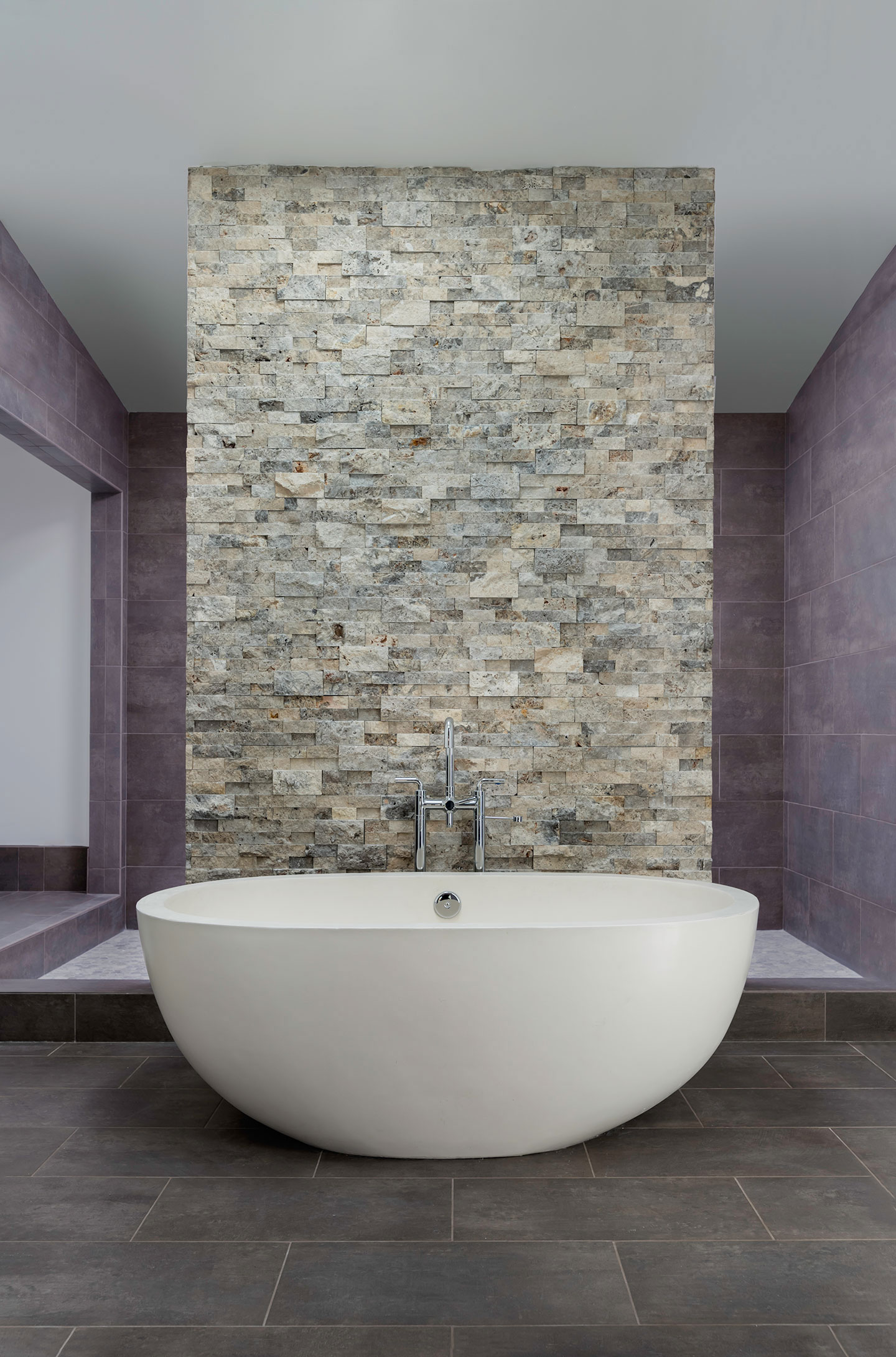 The image shows a modern bathroom interior with a large white freestanding bathtub set against a backdrop of a wall featuring a vertical arrangement of natural stone tiles, which are complemented by a dark-colored floor and a purple accent wall on the left side. Above the bathtub, there is a horizontal light fixture that spans the length of the tub.