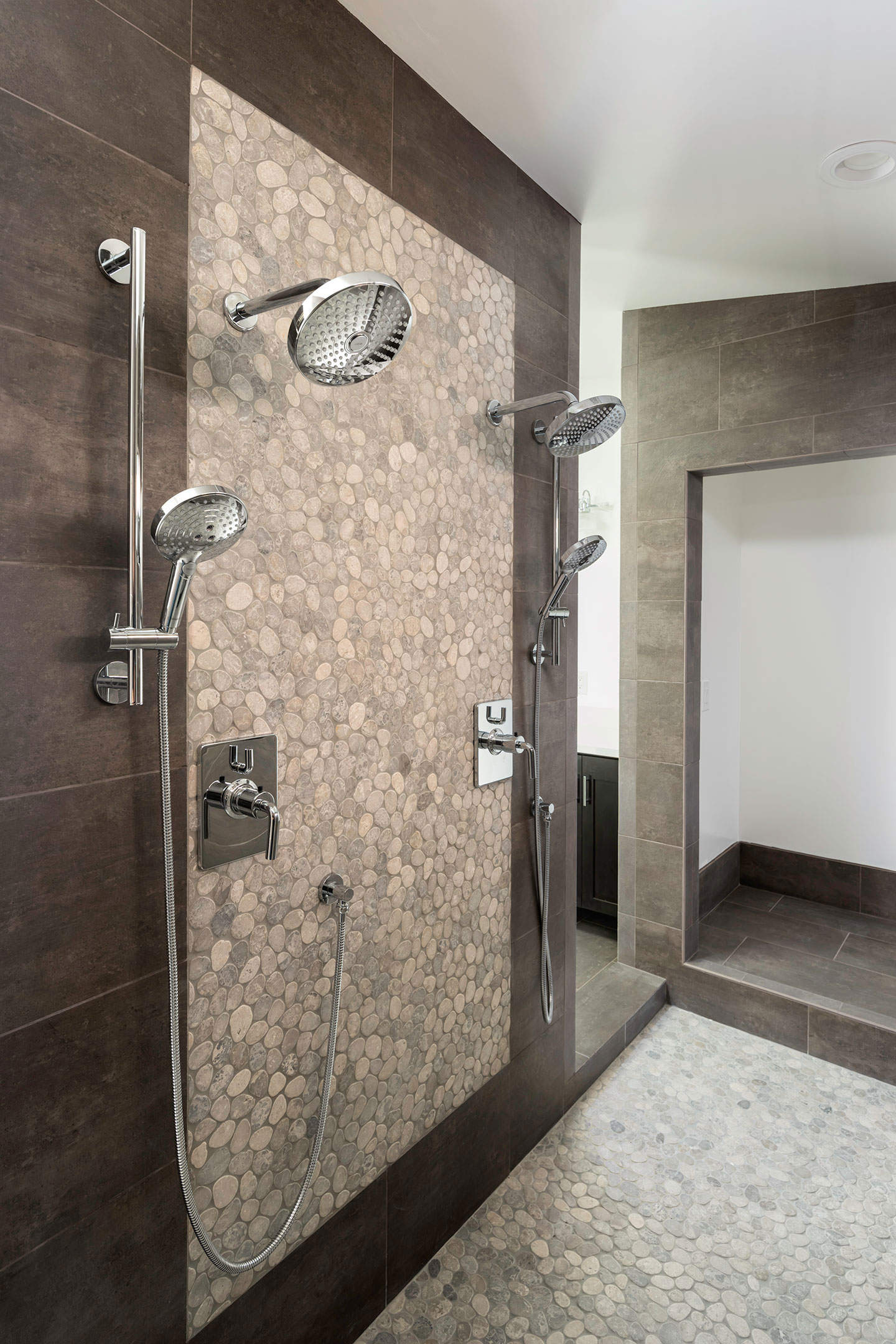 The image shows a modern bathroom interior with a tiled shower area featuring multiple shower heads, a glass door, and a bench seat, alongside a shower stall with a walk-in design and brown tiles on the wall.