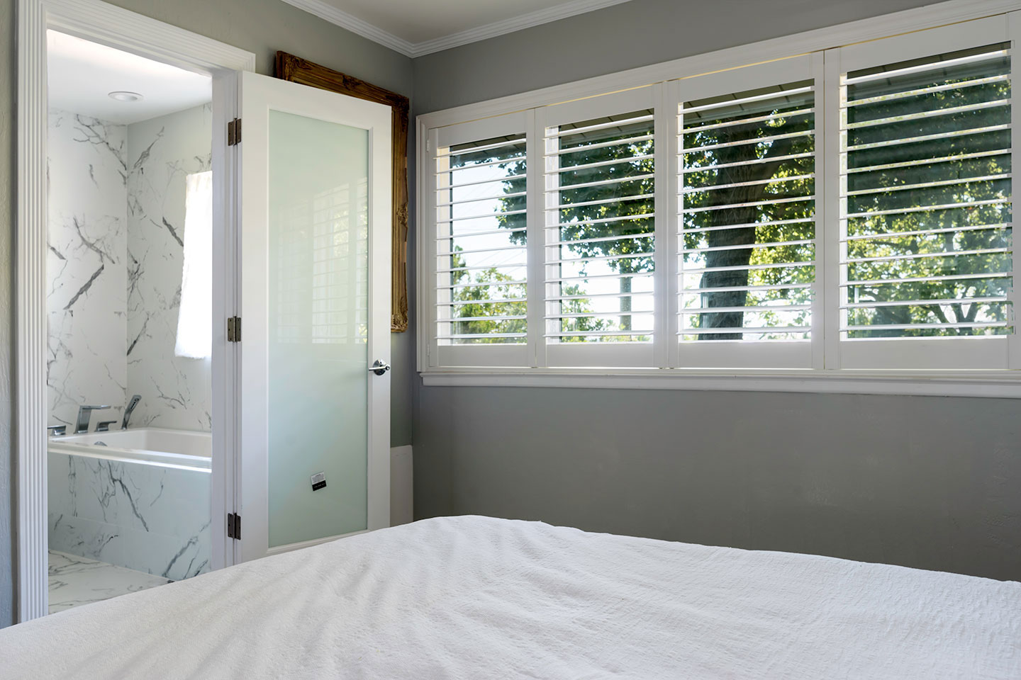 The image shows a modern bedroom with a large window allowing natural light into the room, which features a bed, a white bathtub, a door, and a bathroom window covered by horizontal blinds.
