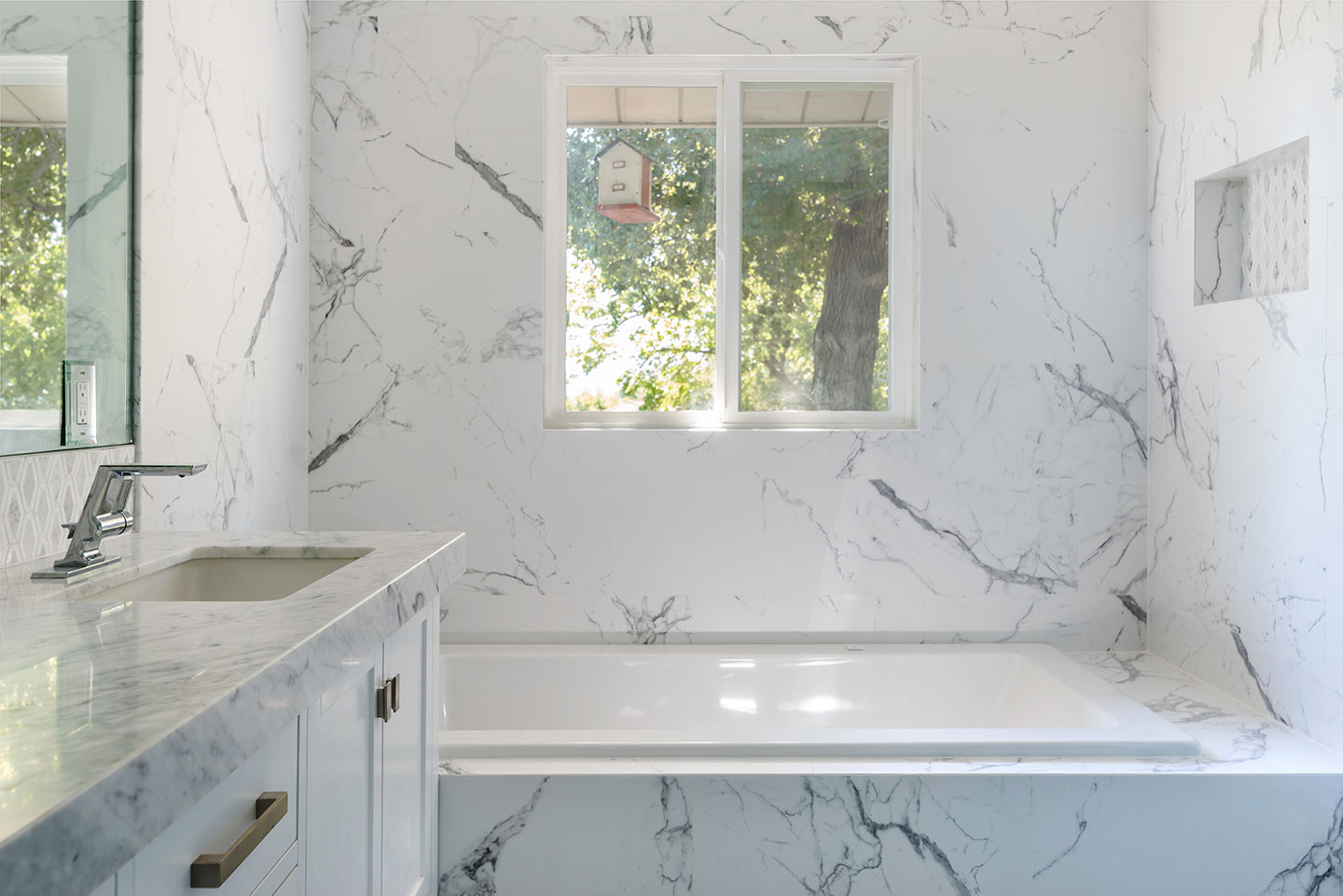 The image shows a modern bathroom with a marble countertop, a large window allowing natural light, and a minimalist design featuring a white color scheme.