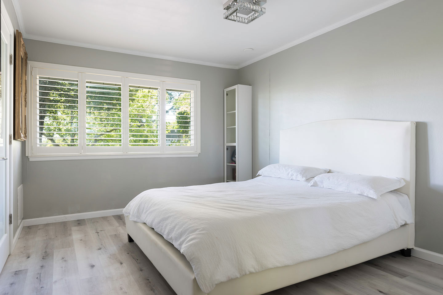 The image shows a neatly made bed in a room with a modern interior design, featuring white walls, light-colored flooring, and a window with horizontal blinds allowing natural light into the space.