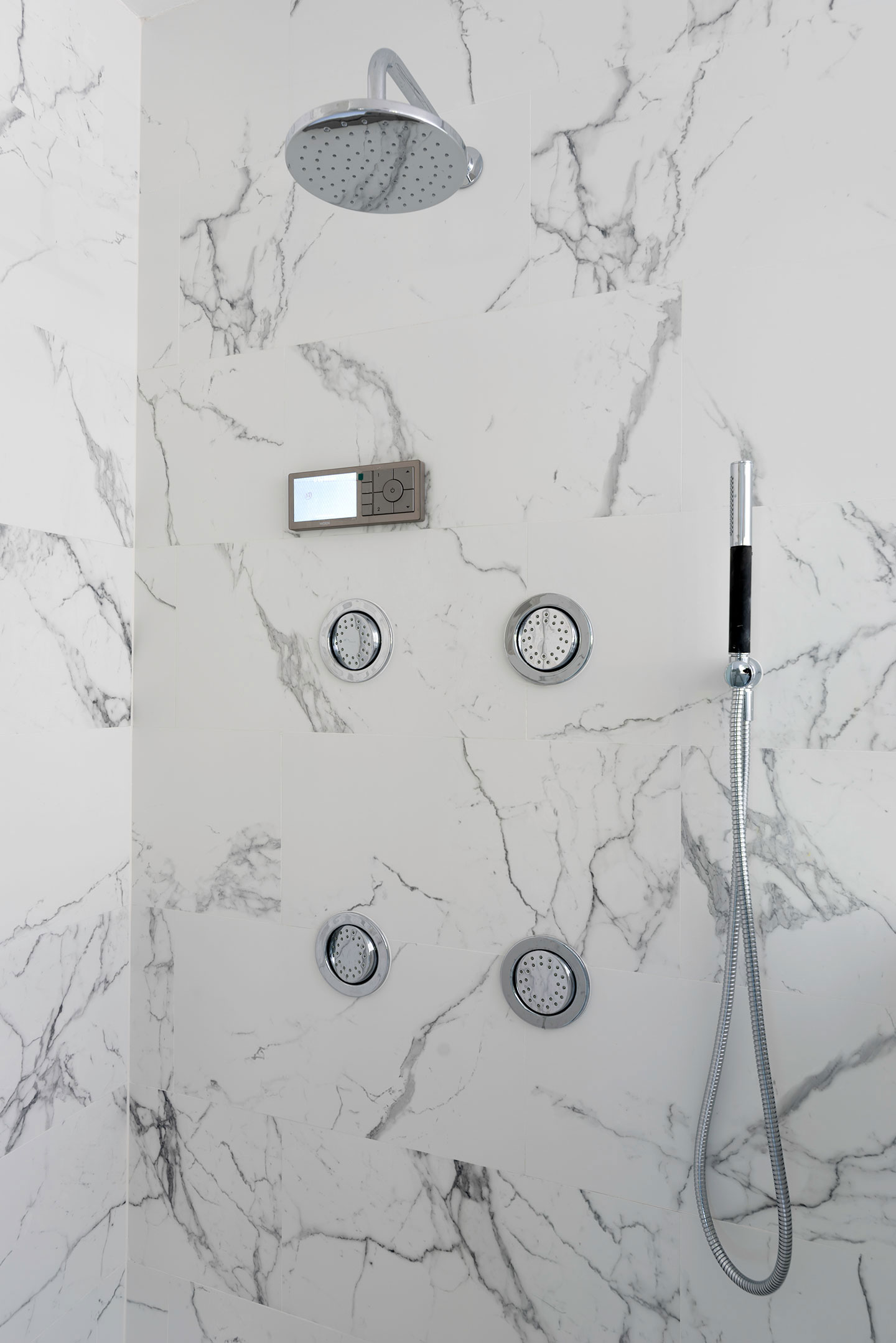 The image shows a modern bathroom interior with marble-patterned wall tiles, a shower head mounted on a wall, a handheld shower head, and various bathroom fixtures including a faucet and multiple shower controls.