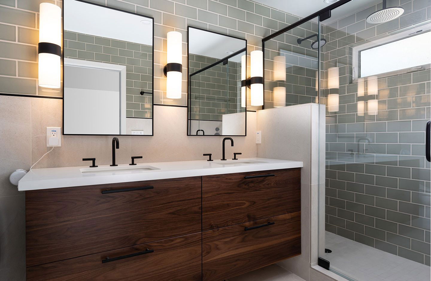 The image shows a bathroom with a modern design featuring a double sink vanity, a large mirror above the sinks, brown cabinetry, and a tiled wall with grey tiles.