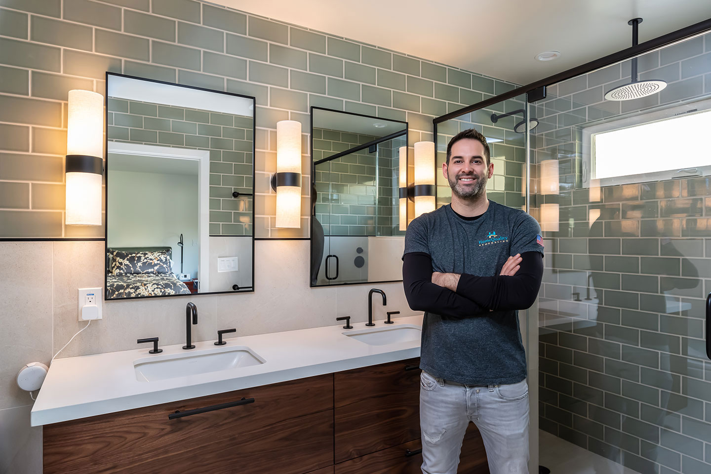 A man stands in front of a bathroom mirror with a modern design featuring metallic accents and dark tiles.