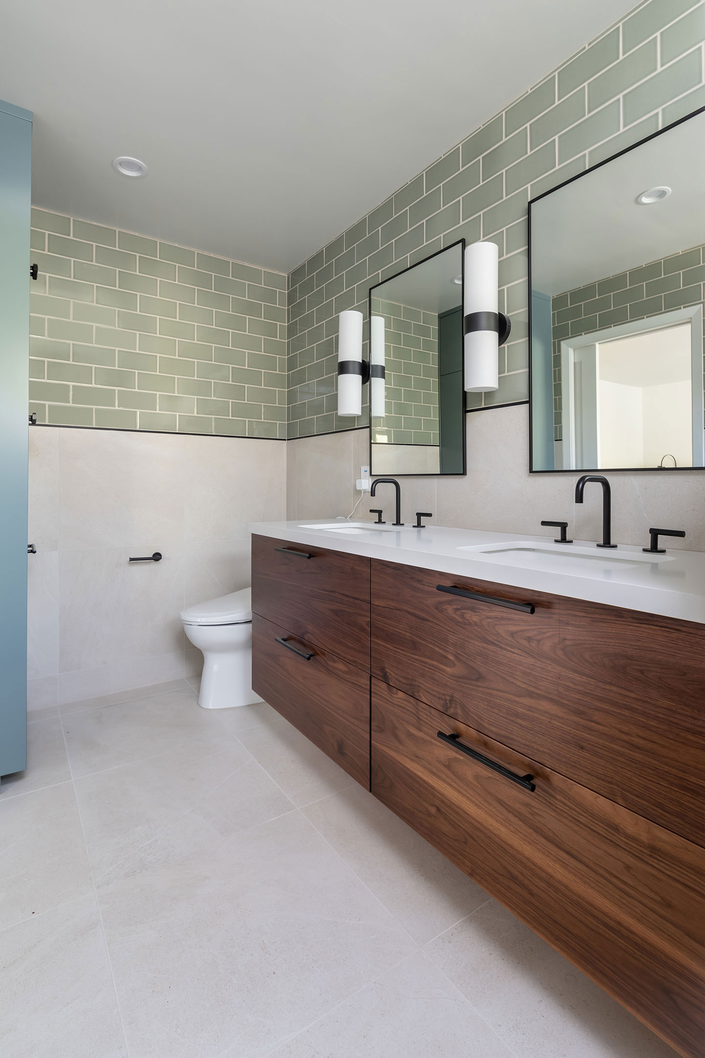 The image shows a modern bathroom with a dark wooden cabinet, a white sink, a mirror above the sink, a toilet, a tiled floor, and green accent tiles on the wall.