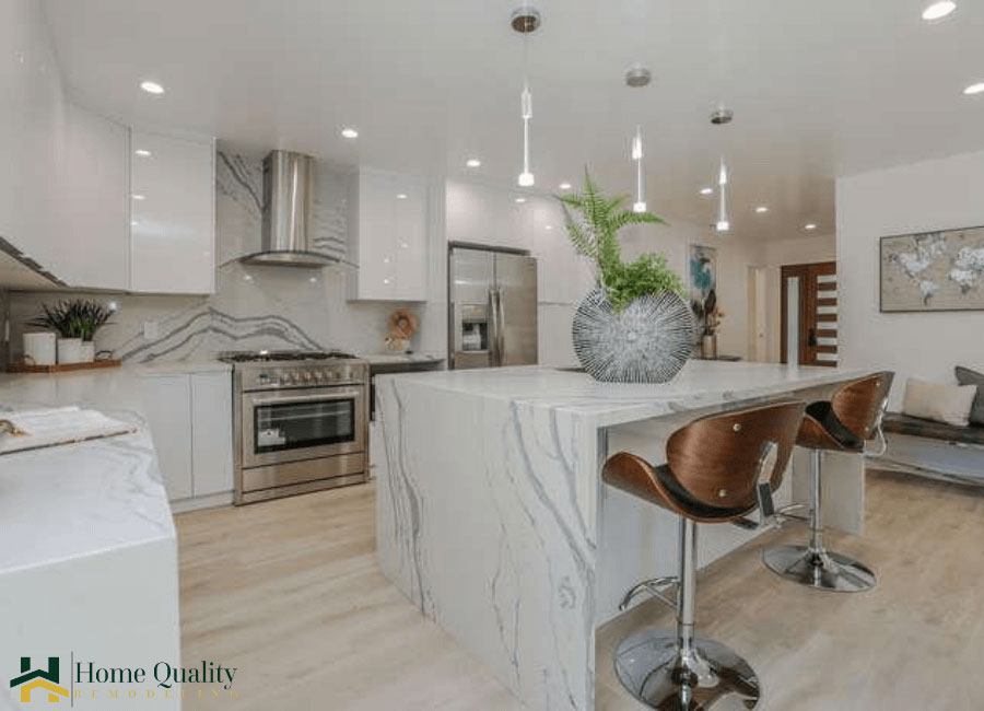 The image shows a modern kitchen interior with marble countertops and a center island, featuring a sink, faucet, and a decorative vase. There are stools around the island, and the room has white walls, a gray cabinetry, stainless steel appliances, and a hardwood floor. The space is well-lit and appears clean and uncluttered.