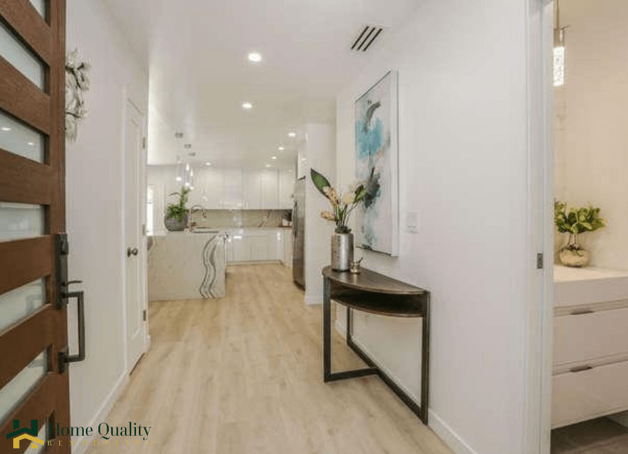 The image shows an interior view of a modern residential kitchen with a clean and minimalist design, featuring white cabinets, stainless steel appliances, a large island countertop, and a hardwood floor.