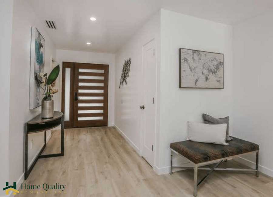 The image shows an interior space with a modern design, featuring a clean, uncluttered hallway leading to a door on the right. The flooring is light-colored wood, and there are decorative elements such as artwork and a plant in the background. The walls are painted in a neutral tone, and natural light appears to be entering from the left side of the image.