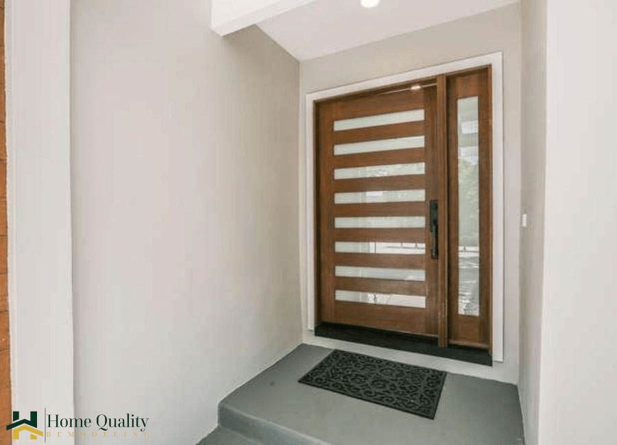 The image shows a modern interior view of a house with a dark wood floor and a white wall. A closed door leads into the room, which features a wooden staircase with glass panels on either side. The door has a glass panel in the center and is framed by a black metal frame.