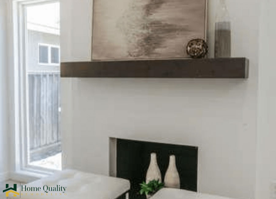 The image shows an interior space featuring a modern wall-mounted shelf with a framed artwork above a white fireplace, which has a black grate. A vase sits on the mantel below the artwork, and there are two small decorative items to the left of the fireplace. The wall is painted in a light color, and the flooring appears to be dark wood or laminate. The overall style suggests a contemporary home setting with neutral tones and minimalist design elements.