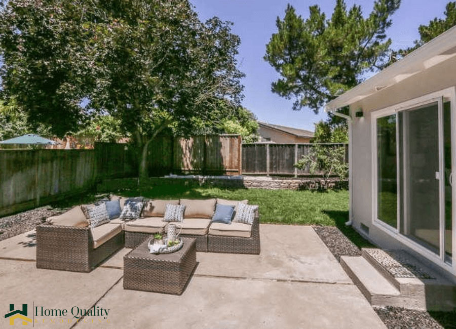 The image shows an outdoor patio area with a large dining table surrounded by chairs, a paved seating area with additional chairs and a coffee table, a lawn, a fence, and a backyard with trees and a clear sky overhead.