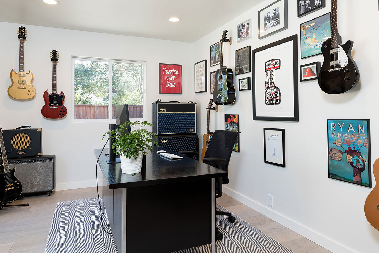A wall covered with various framed items, including posters and photos, showcasing a collection of guitars and music-related memorabilia, set against a backdrop of a room with musical equipment and a desk, suggesting a home office or studio dedicated to music enthusiasts.