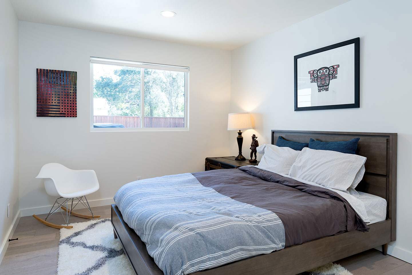 The image shows a neatly arranged bedroom with a large bed, nightstands, a dresser, a chair, a framed picture on the wall, and a window with blinds partially drawn.