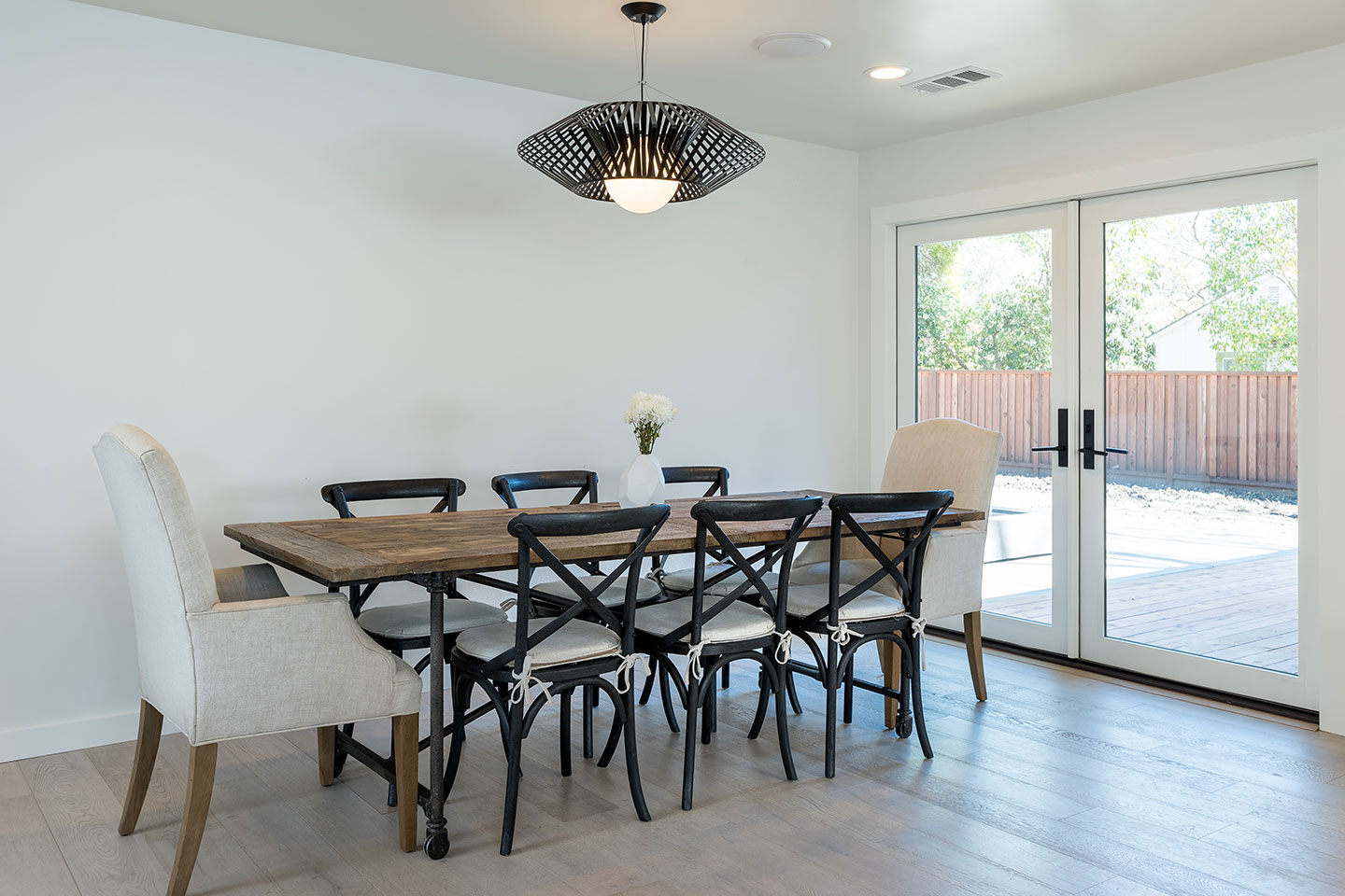 The image depicts a modern dining room with a dark wood floor, a white wall, a ceiling light fixture, a large dining table surrounded by eight chairs, and a sliding glass door leading outside.