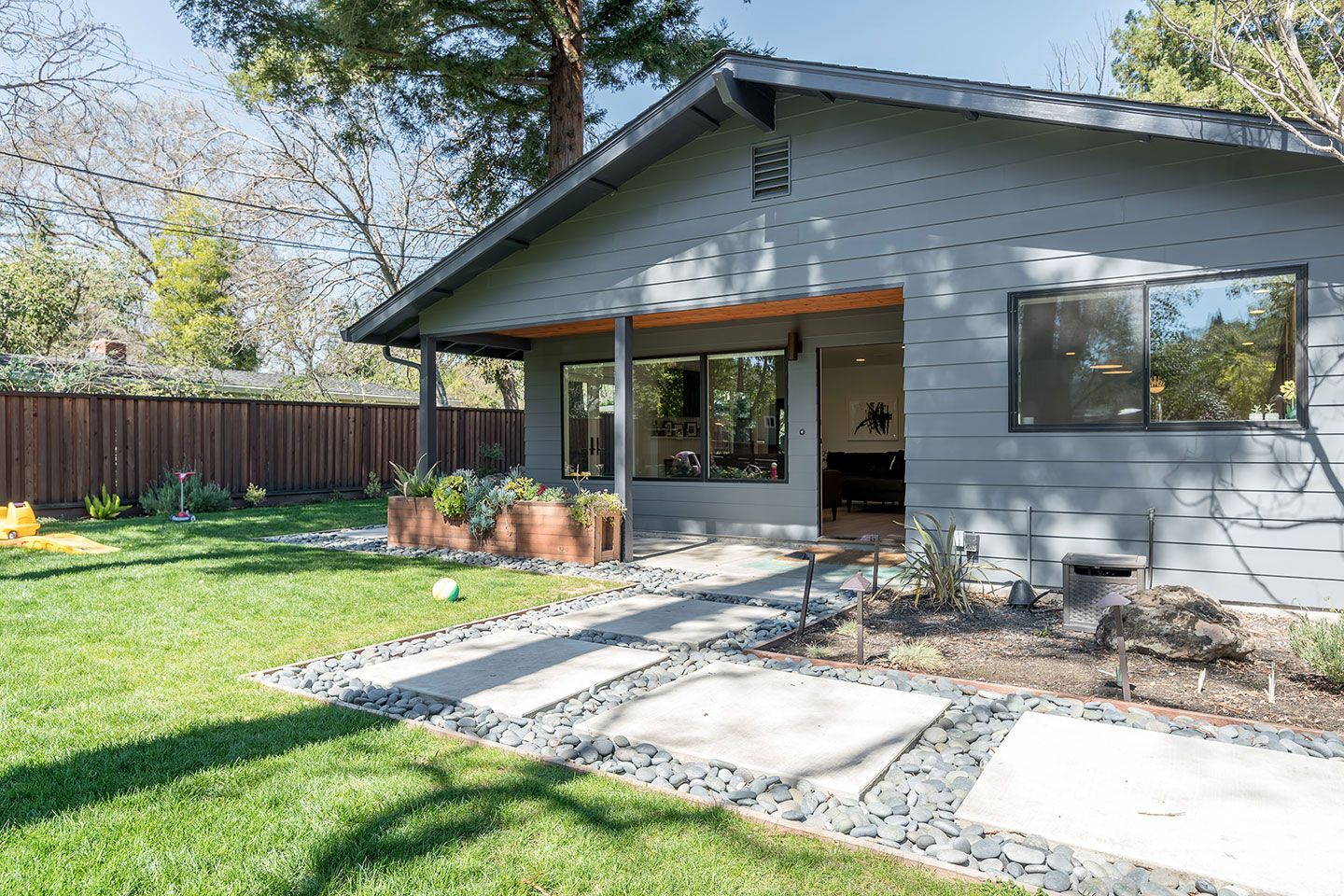 The image shows a modern single-story house with a gray exterior, a dark roof, and a large deck area. The house features multiple windows, a prominent front door, and is surrounded by a well-maintained yard with paving stones leading to the entrance.