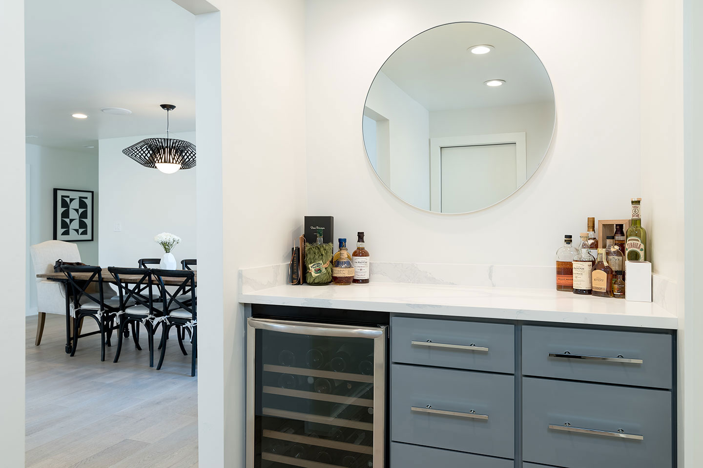 The image shows a modern kitchen interior with a white color scheme, featuring a large island countertop, a stainless steel sink, a mirror above the counter, a refrigerator, and a dining table set up for four people.