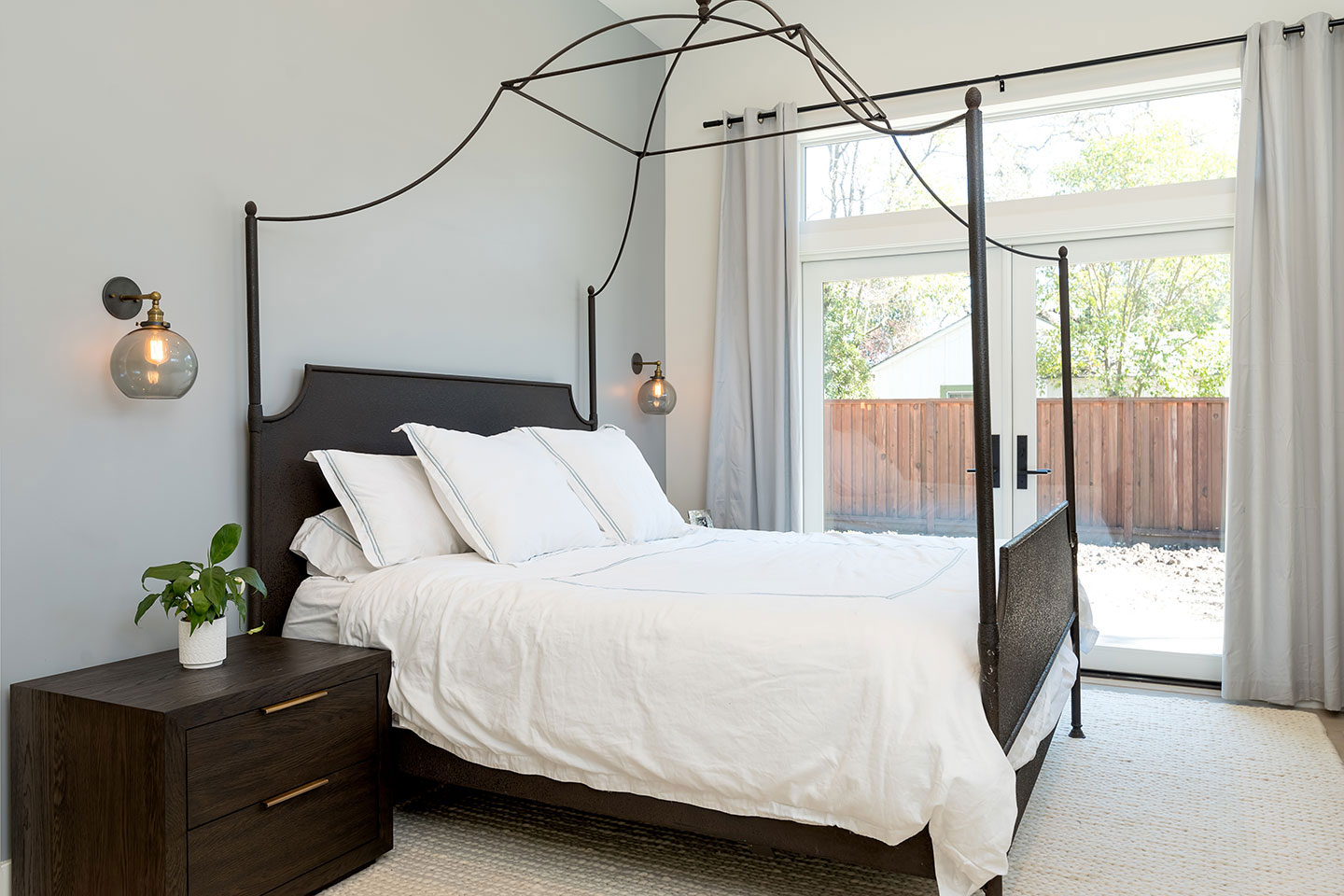 A modern bedroom with a large bed, wooden headboard, black metal frame, white linens, wall-mounted lighting, and a window with curtains.