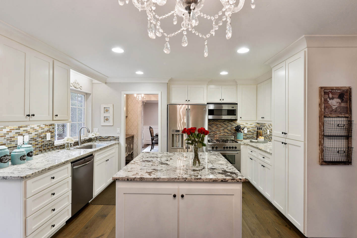 The image depicts a modern kitchen interior with white cabinets, marble countertops, stainless steel appliances, a center island with a sink, a chandelier hanging from the ceiling, and various decorative elements such as a vase, a clock, and a framed picture.