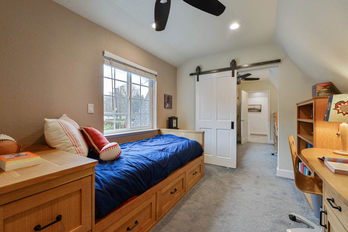 The image shows a bedroom with a neatly made bed, a desk area with a chair, a bookshelf, and various items on top, a closet with an open door revealing hanging clothes, a ceiling fan, and a window allowing natural light into the room.