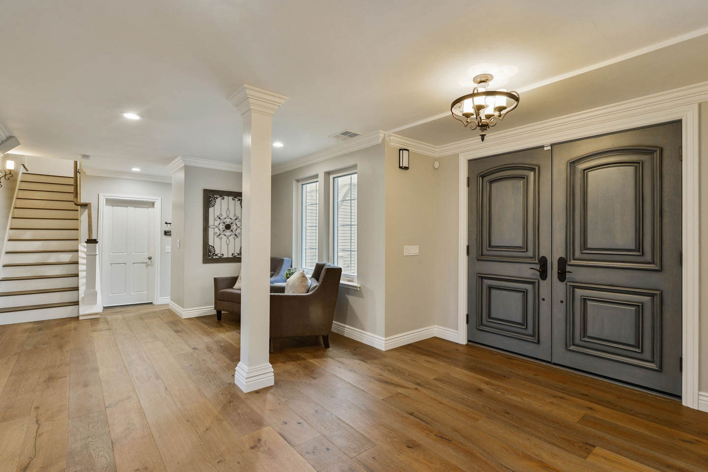 This image shows a well-lit interior hallway with hardwood floors, a modern staircase with a dark wood handrail, and doors on either side leading to different rooms. The walls are painted white, and there s a chandelier hanging from the ceiling.