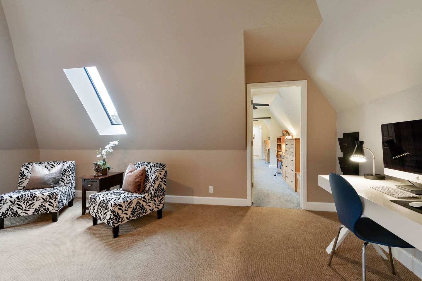 This is a photograph of an interior space, likely a living room, with a sloped ceiling and a modern design featuring furniture such as a sofa, chairs, and a desk, set against a neutral color palette with a large window allowing natural light to enter.