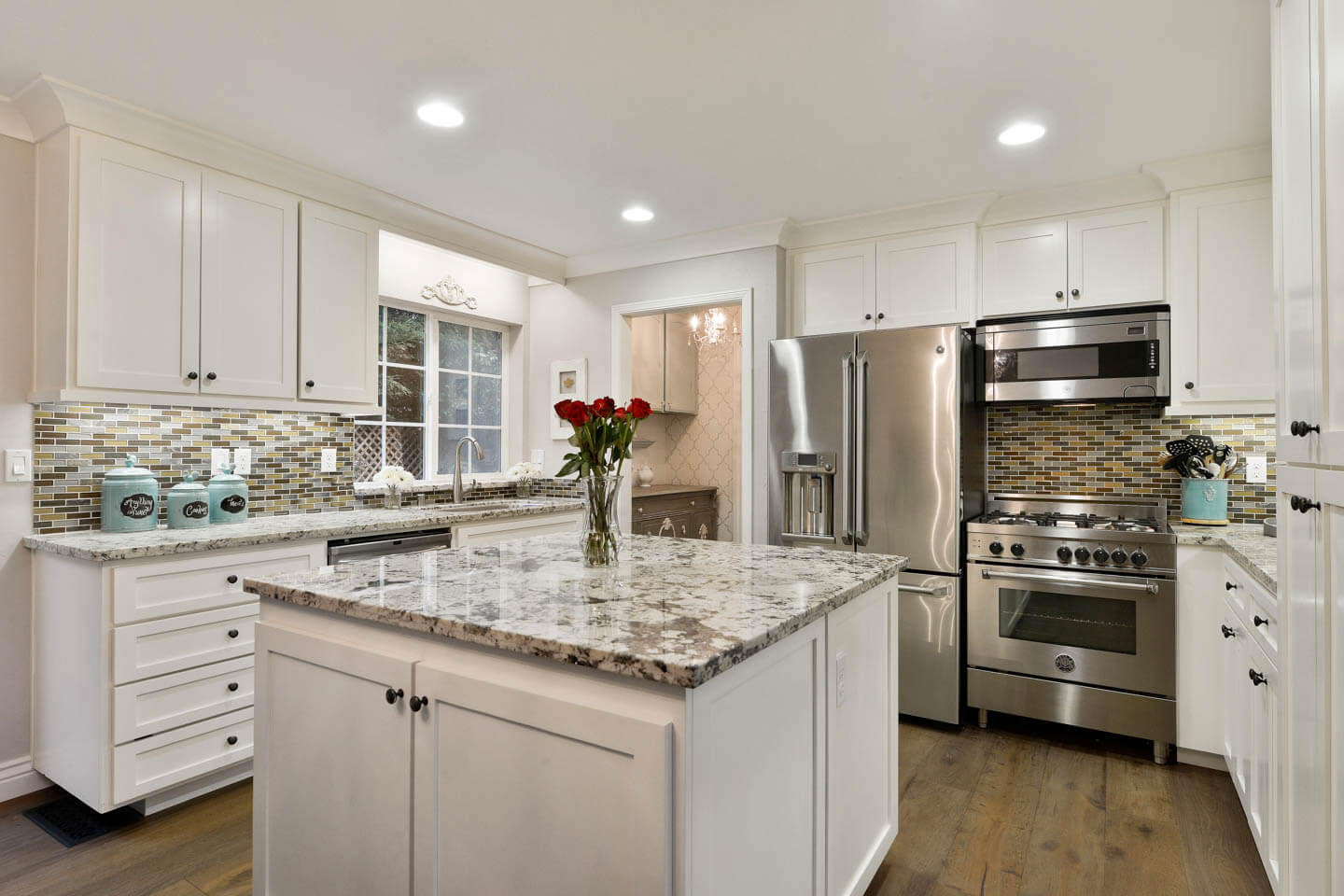 The image shows a modern kitchen with white cabinets, marble countertops, stainless steel appliances including a refrigerator, oven, and sink, a center island with a built-in stove, and a backsplash featuring a geometric pattern.