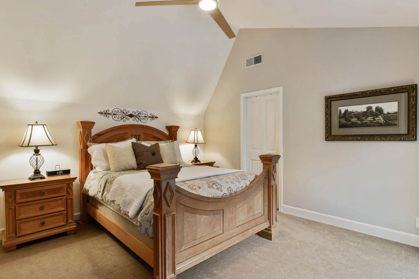The image shows a well-furnished bedroom with a large wooden bed frame, a neatly made bed with multiple pillows, a nightstand with a lamp on one side, a framed picture on the wall above the bed, and a ceiling fan with lights.