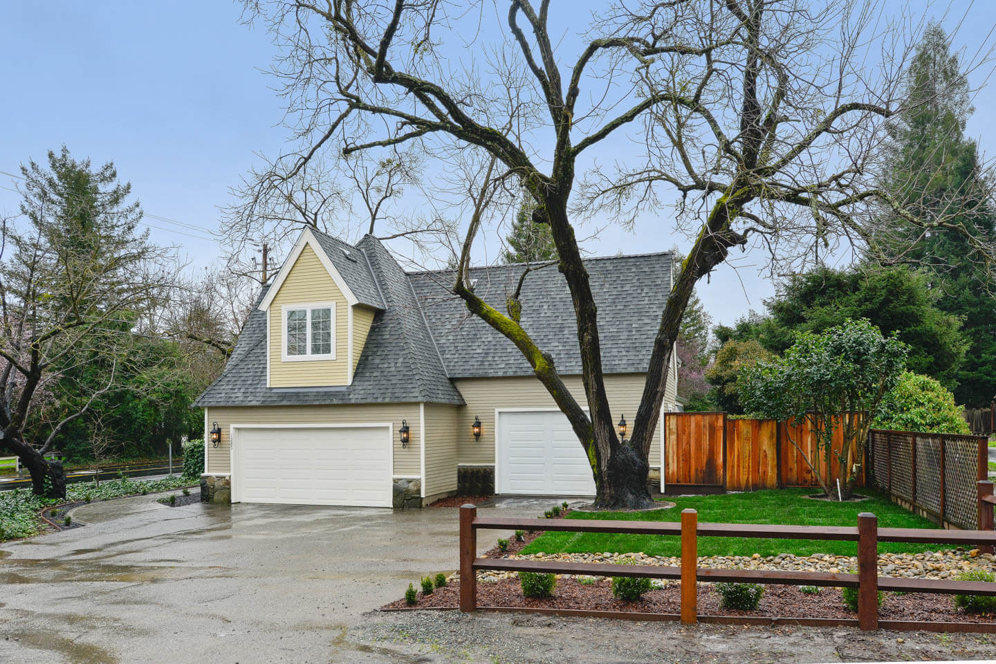 The image shows a two-story house with a garage door, set against a backdrop of trees and a clear sky.