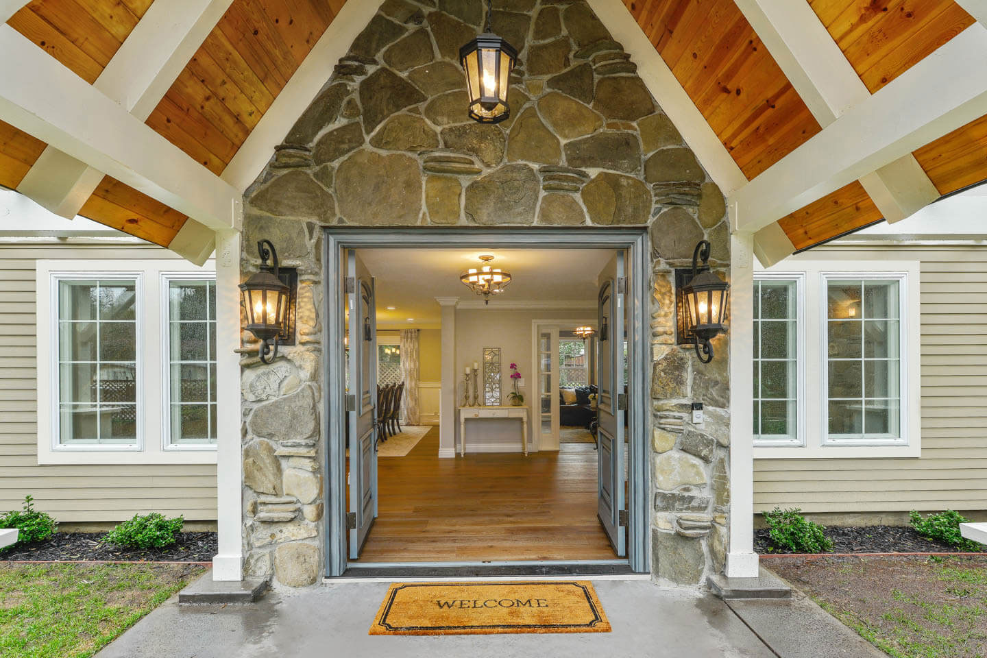 The image shows a large two-story house with a prominent front entrance featuring a covered porch, stone pillars, and a gabled roof with exposed wooden beams. The exterior walls of the house are painted in shades of white and gray, and there s a garage door on the left side of the house.