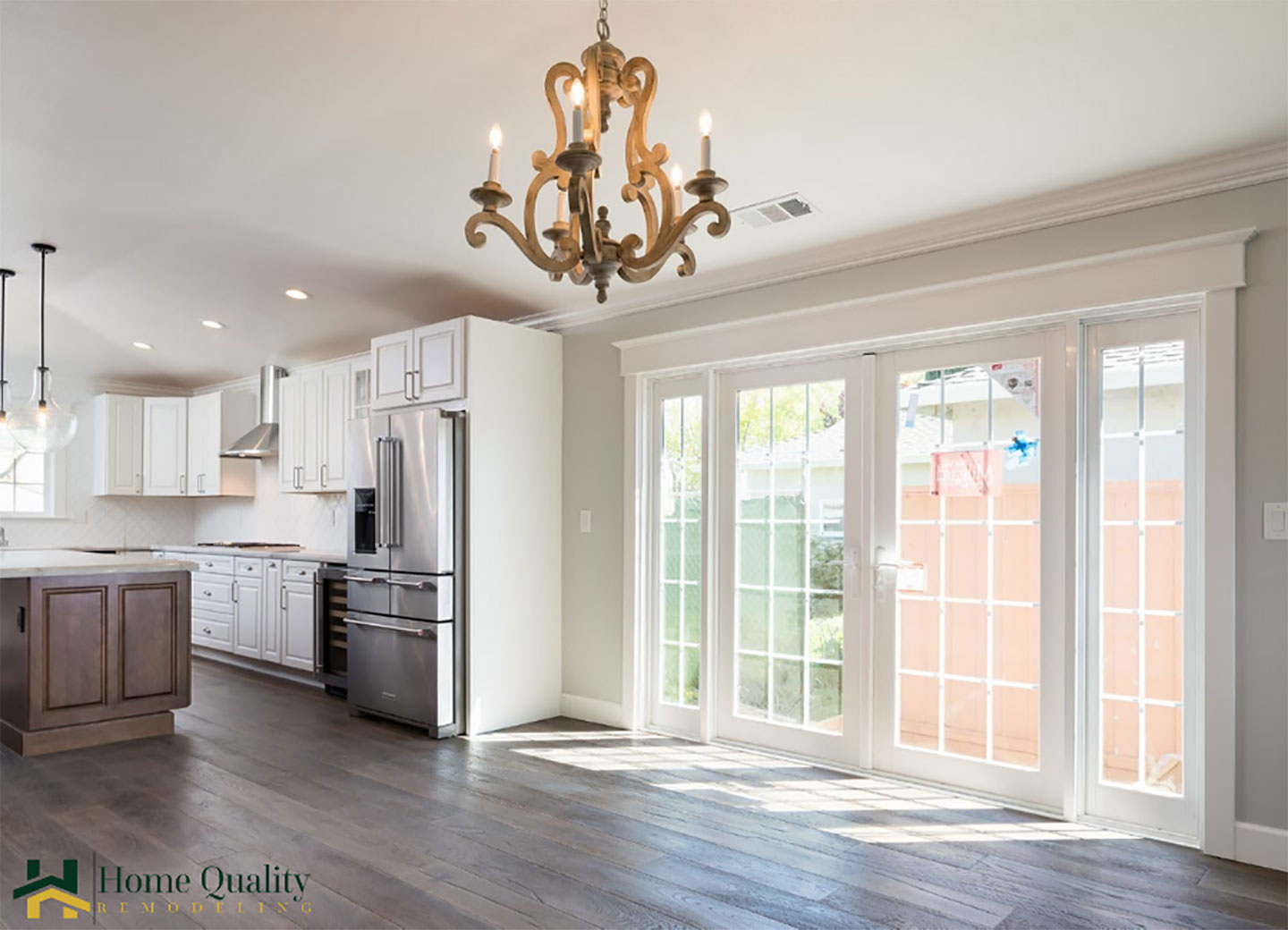 A well-lit kitchen with modern appliances, white cabinets, and hardwood floors, featuring an island with a sink, a large window allowing natural light, and a chandelier hanging from the ceiling.