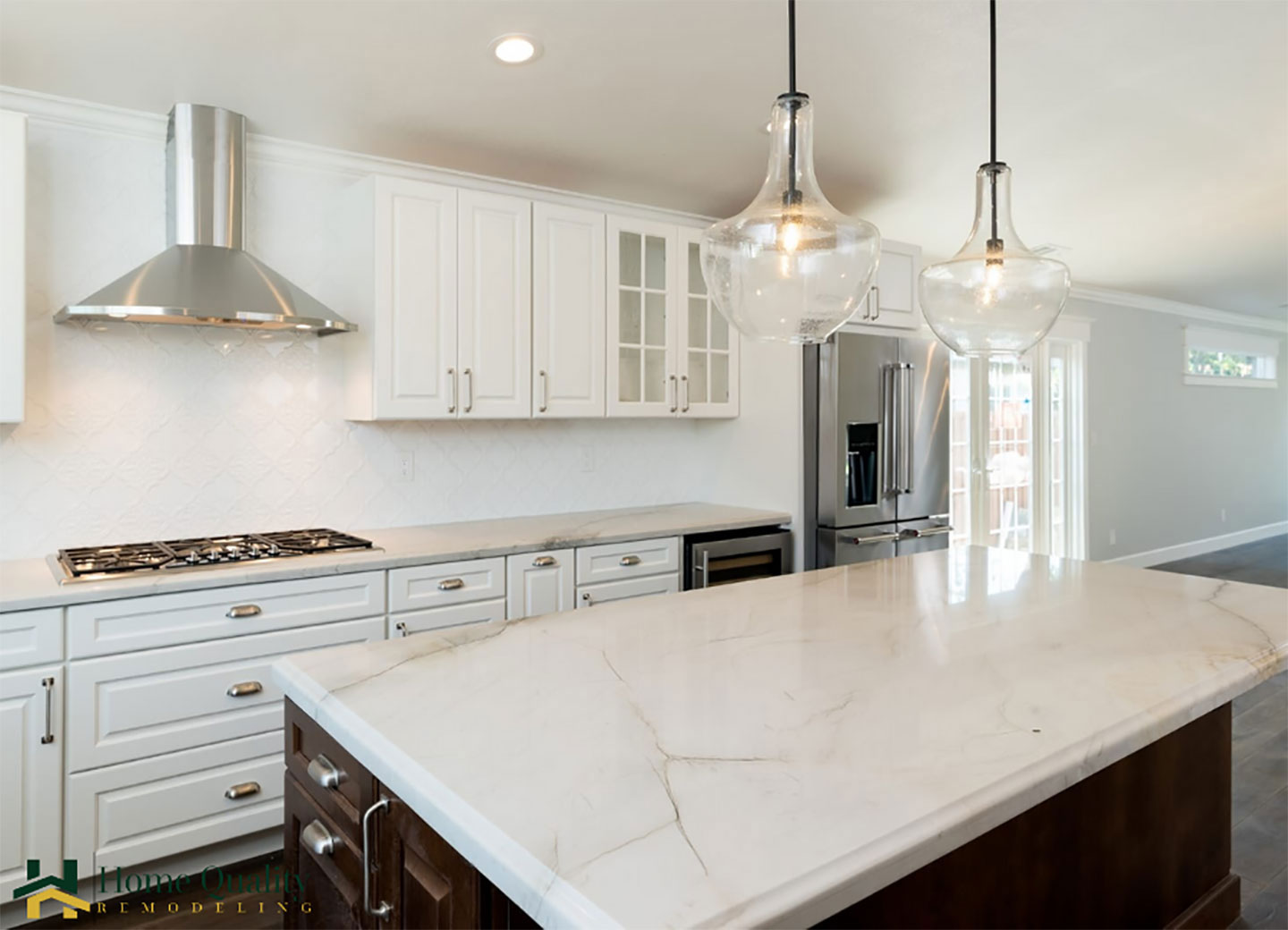 A modern kitchen with white cabinets, marble countertops, stainless steel appliances, pendant lighting, and a large island in the center.