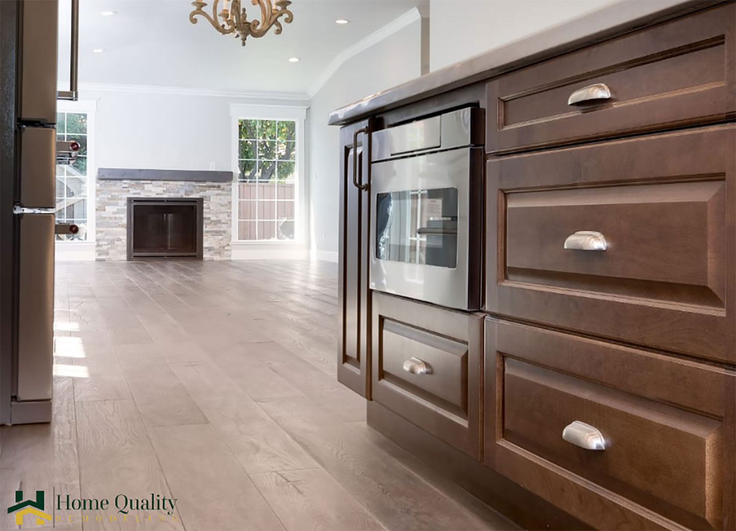 The image shows a modern kitchen interior with a prominent brown cabinetry set against a white wall, featuring stainless steel appliances including an oven and refrigerator, a large island with a sink, and a hardwood floor.