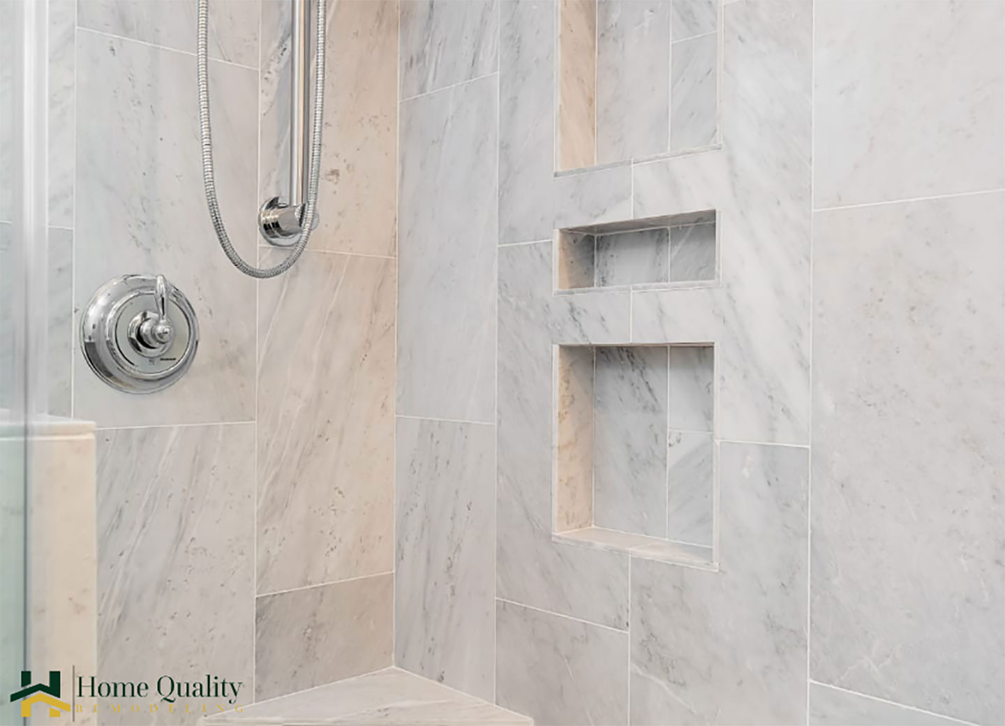 The image depicts a modern bathroom with a marble-tiled shower enclosure featuring a walk-in design, a built-in bench, and a glass door. A chrome showerhead is visible, along with white fixtures and a dark gray floor. The shower is set against a white wall with recessed shelving on the right side.