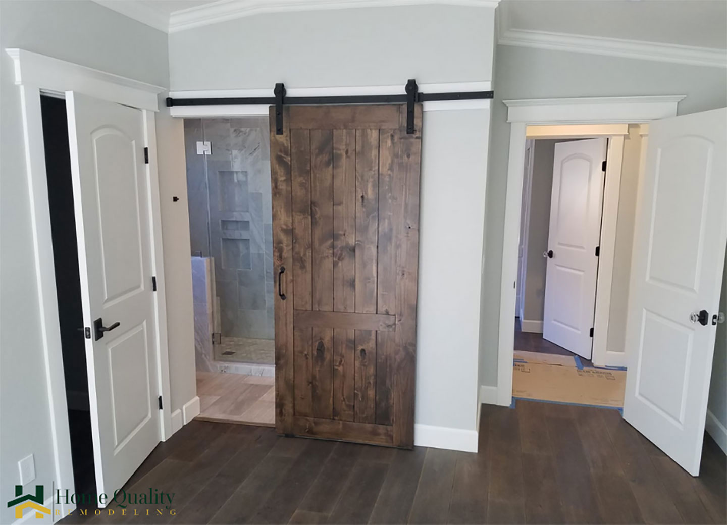 This is an interior view of a room with a prominent wooden door featuring a horizontal slat design, set against a textured wall with a darker tone. The floor appears to be a light-colored hardwood, and there s a partial view of another door on the right side with a similar style.