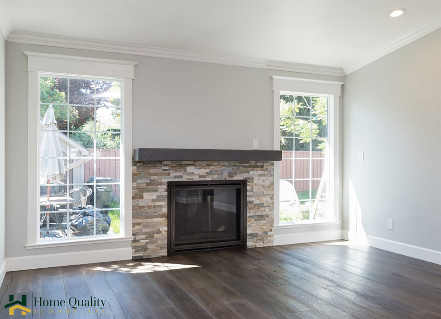 The image shows an interior view of a living room with a modern fireplace, stone veneer mantel, hardwood flooring, and large windows with white frames.