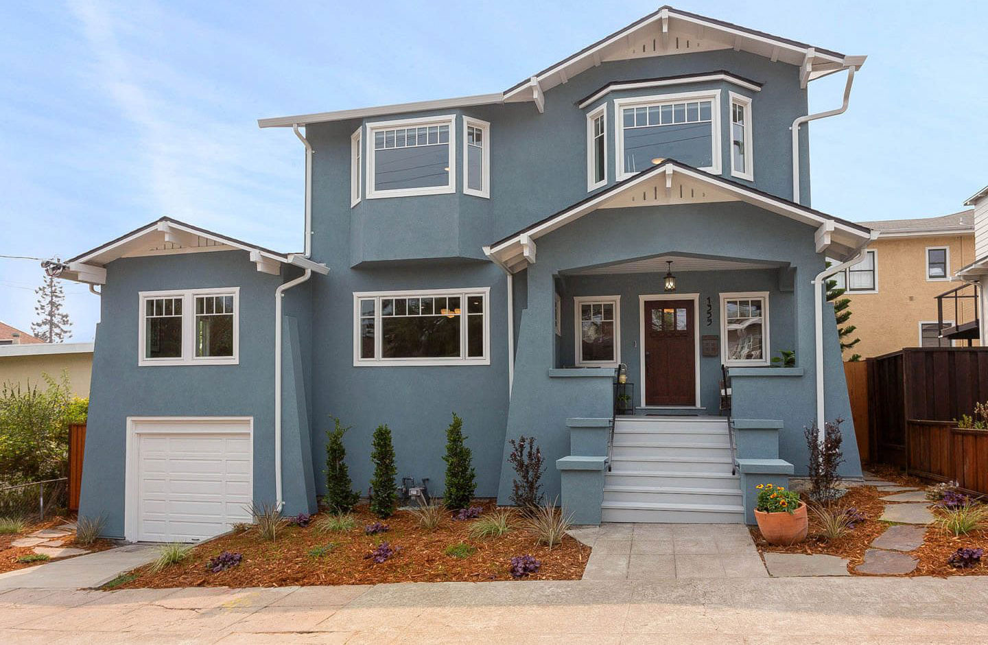 The image shows a two-story house with a white exterior, multiple windows, and a front porch. It has a garage door on the left side and is situated on a residential street with a clear sky above.