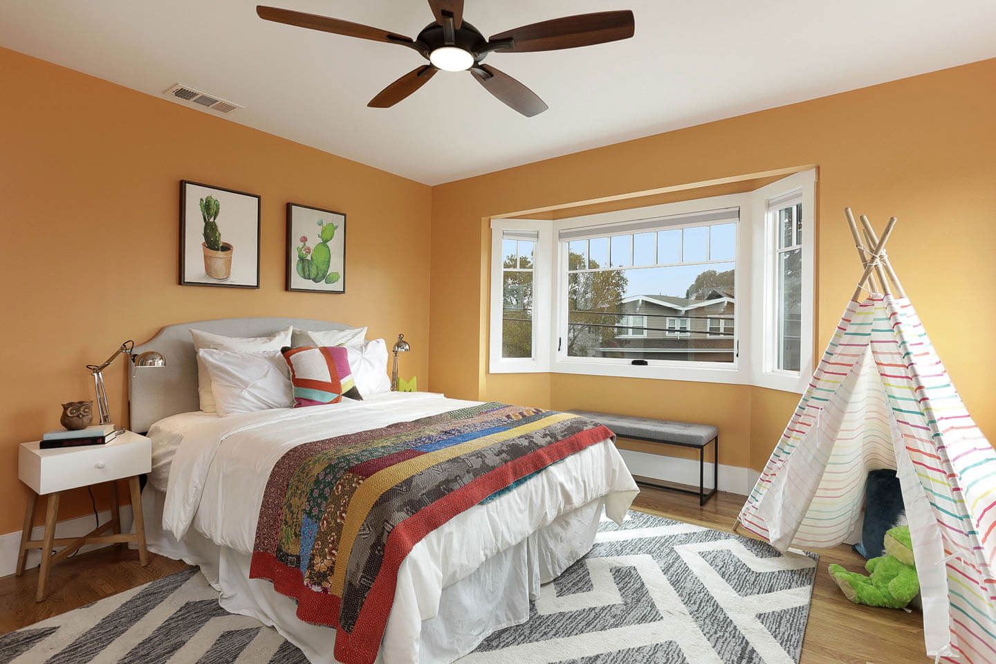 The image shows a well-decorated bedroom with a large bed, colorful bedding, a wooden dresser, a teepee tent, a desk with a lamp, and a window with blinds partially drawn up.