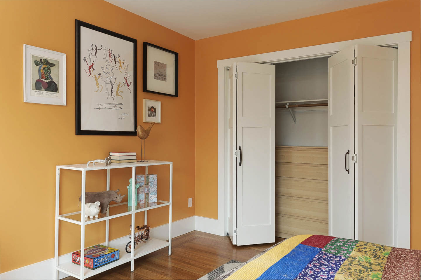 The image shows a room with a bright yellow wall, a closet with white doors, a small bookcase next to the closet, a framed picture on the wall, a shelf above the door frame, and a bed in the background with a colorful quilt.