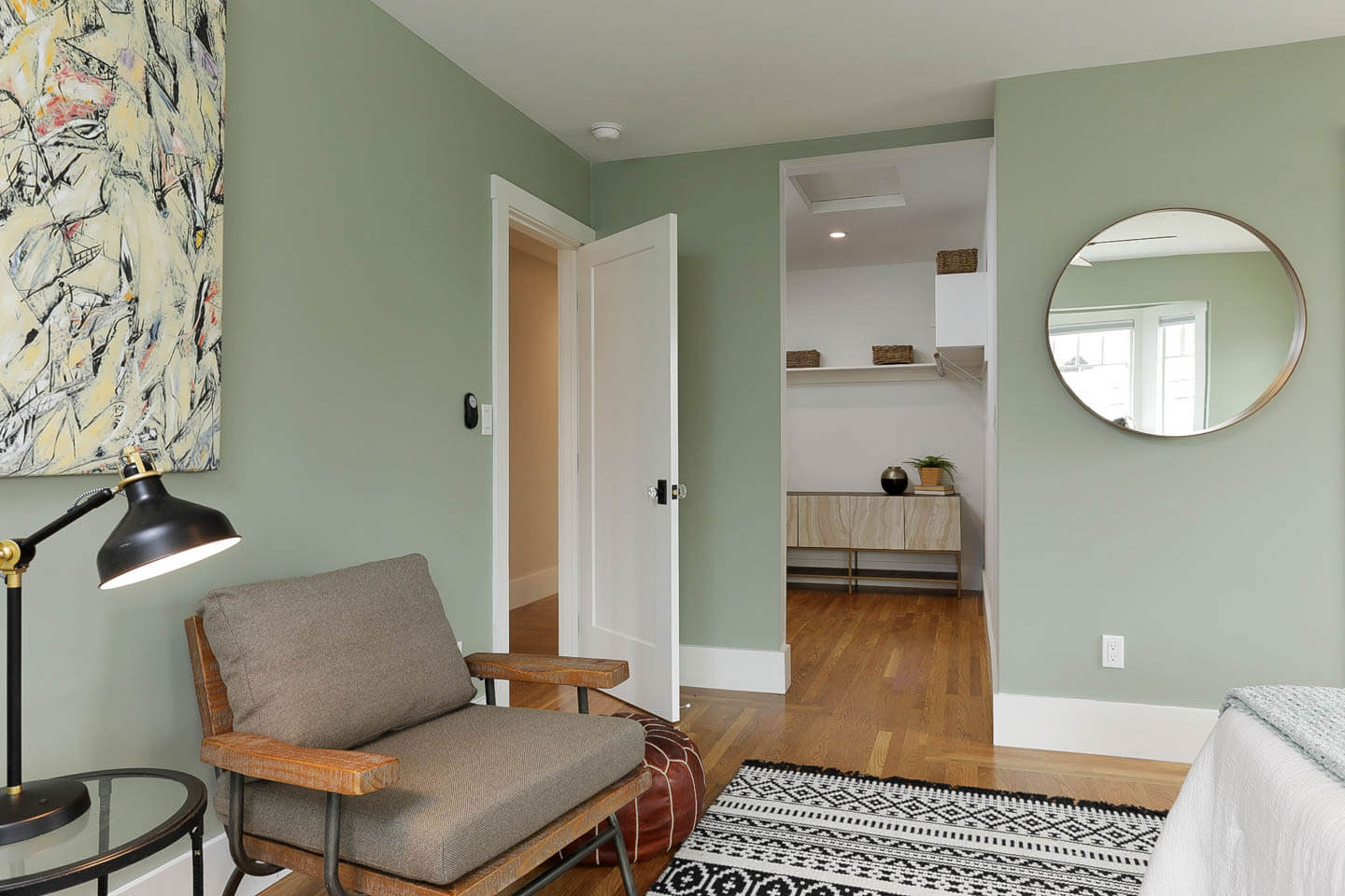 The image shows a modern interior space with a minimalist design, featuring a bedroom area with a bed, nightstand, and wall-mounted shelves, and an adjacent living room area with a sofa, chair, side table, and framed artwork on the walls.
