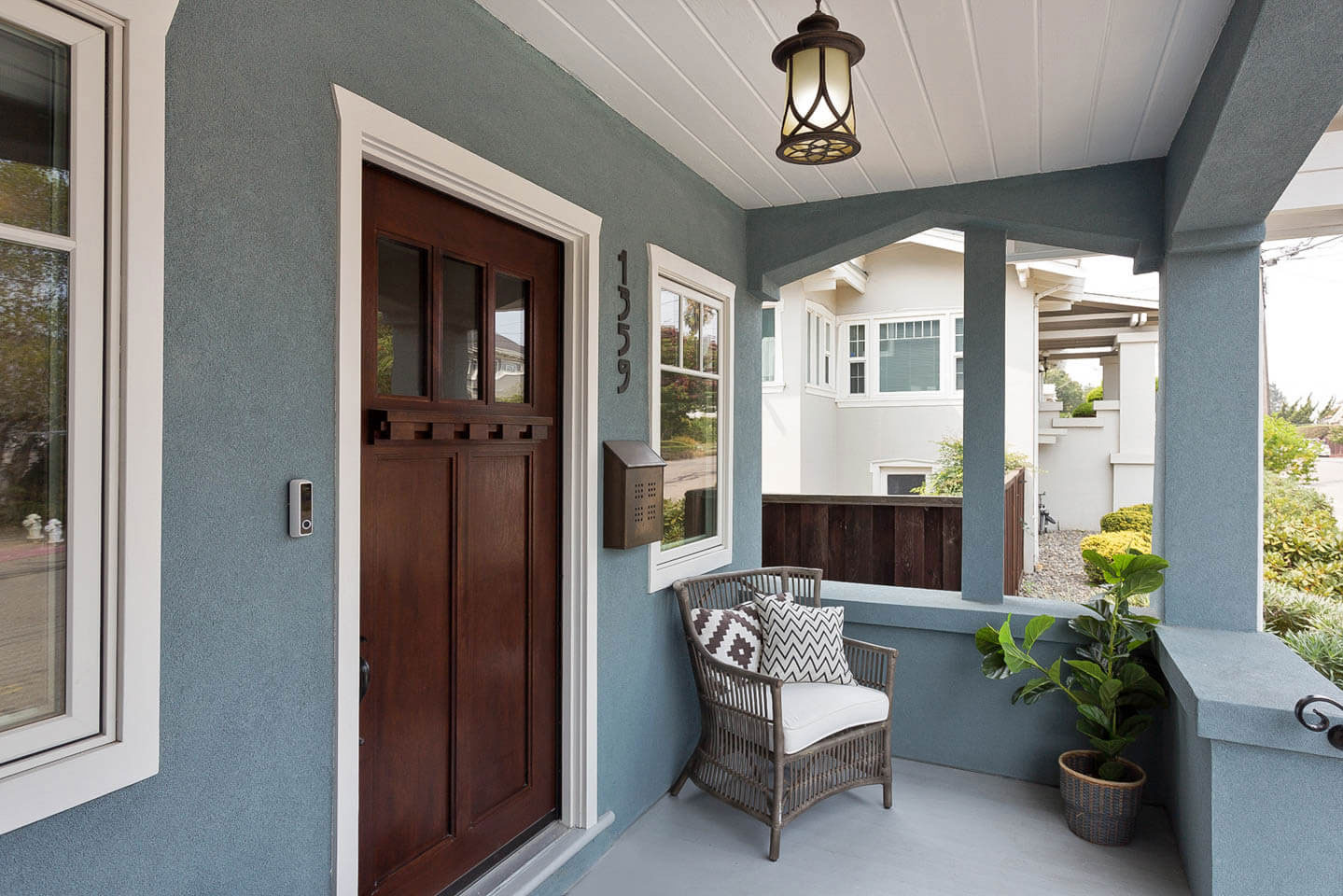 The image shows a front porch with a door on the right side, featuring a blue exterior wall, white trim around the windows and doors, and a green house number.