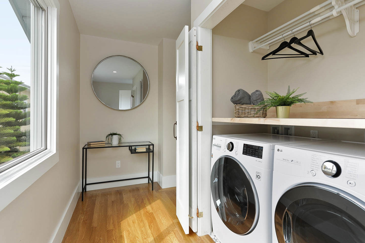 This is an interior photograph of a small, well-lit room with hardwood floors, featuring a laundry machine, a mirror, and a window with natural light.