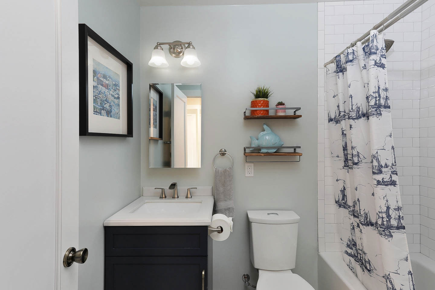 The image shows a small bathroom with a sink, toilet, and shower curtain featuring a nautical design.