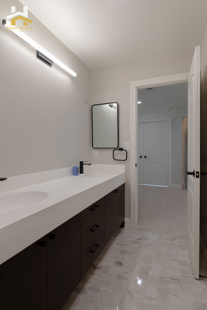 The image shows a modern bathroom interior with a sink, mirror, and vanity area, featuring a minimalist design with dark wood accents and white countertops.