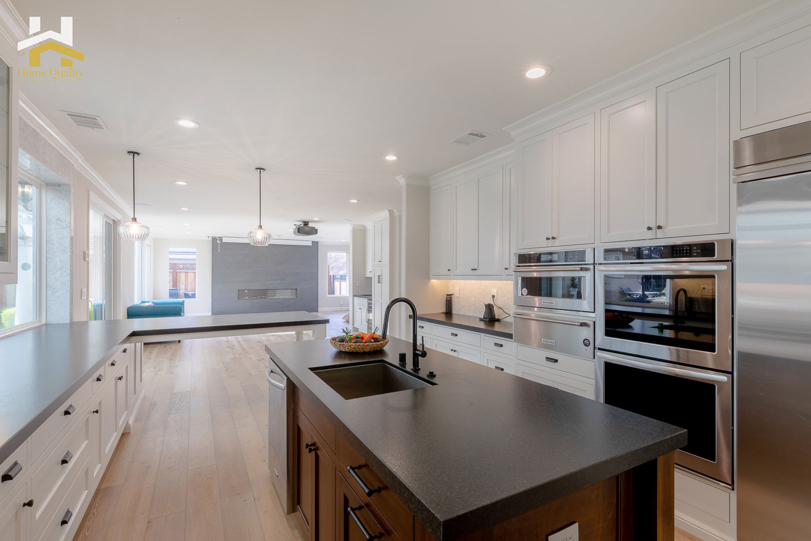 The image shows a modern kitchen interior with white cabinets, black countertops, stainless steel appliances, and a central island with a cooktop.