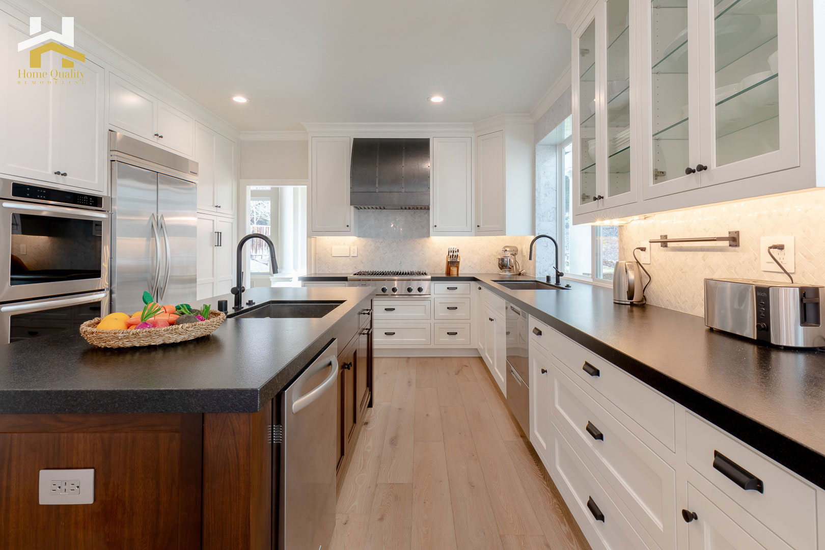 The image depicts a modern kitchen interior with white cabinetry, dark countertops, stainless steel appliances, black flooring, and a central island with a sink. The kitchen features a color scheme of neutral tones with accents of black and silver.