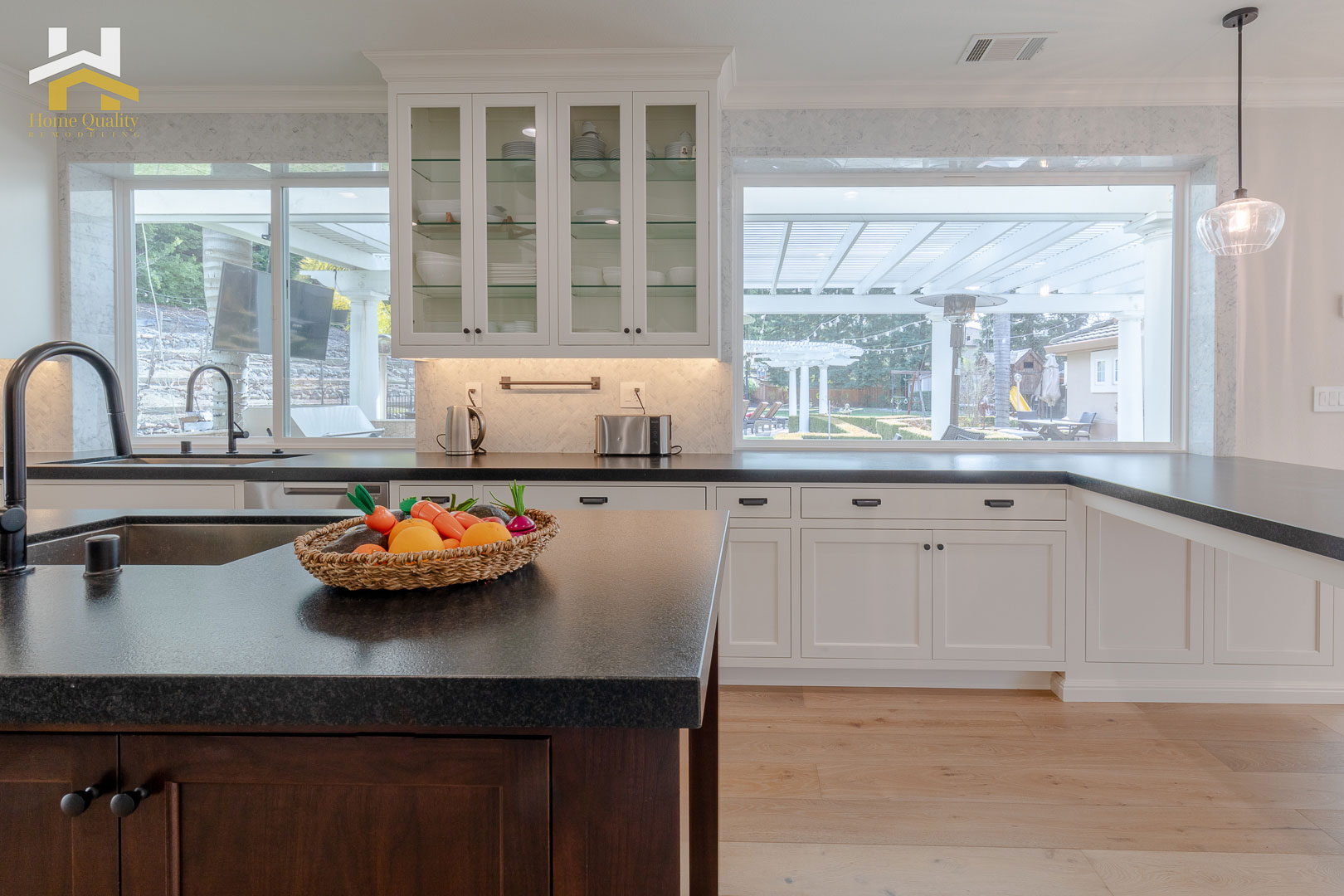 The image features a modern kitchen interior with light-colored cabinets, a dark countertop, stainless steel appliances, a center island with a sink, and a large window allowing natural light into the space.