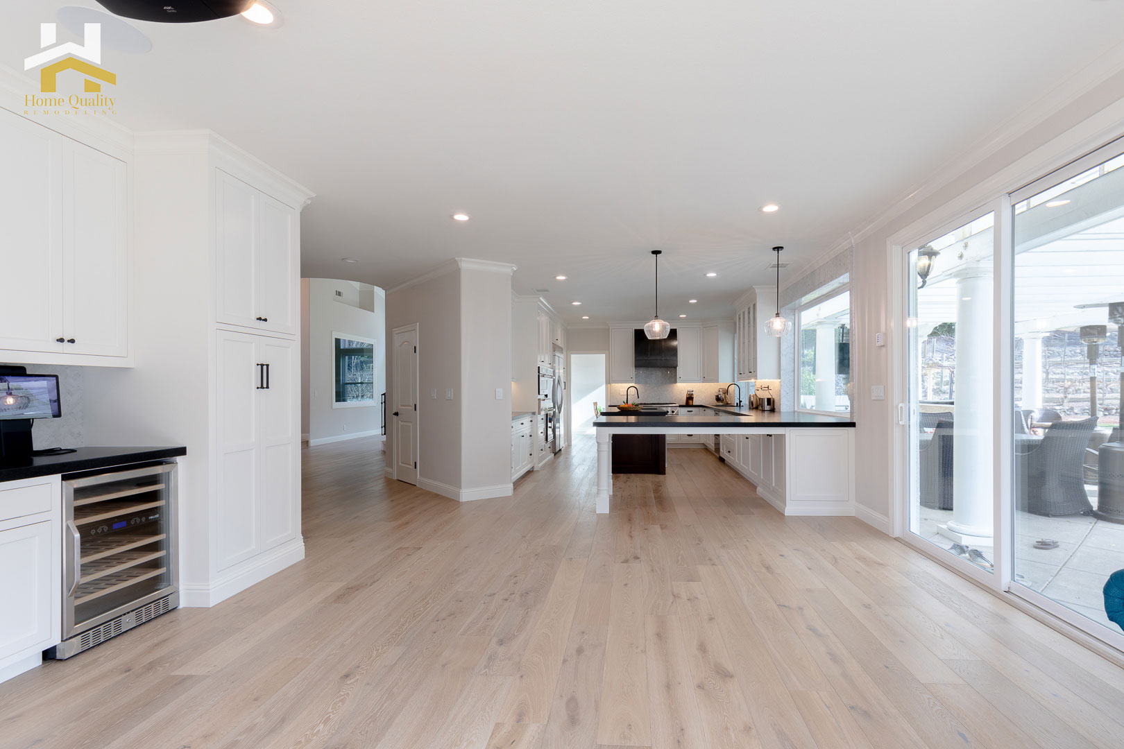 The image shows an interior view of a modern kitchen with white cabinets, hardwood flooring, and large windows allowing natural light into the room.