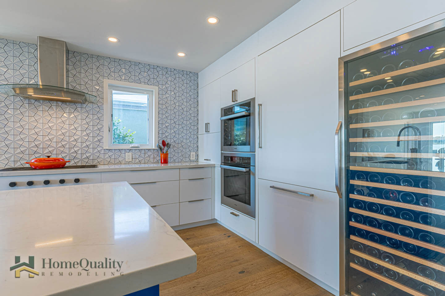 The image shows a modern kitchen interior with white cabinets, stainless steel appliances, a wine rack, and a patterned backsplash.